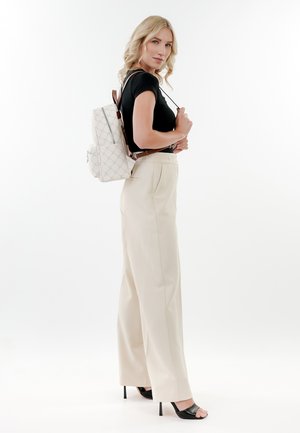 Weißer Rucksack mit geometrischem Muster und braunen Akzenten, lässig über einem schwarzen Oberteil und cremefarbenen, weit geschnittenen Hosen getragen, dazu offene schwarze High Heels.