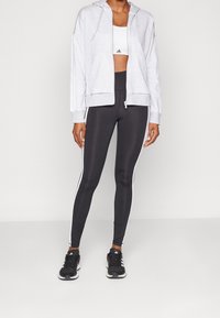 Grå zip-up hoodie med dragsko, tillsammans med högmidjade svarta leggings med vita sidostrecker. Avslutad med svarta träningsskor.