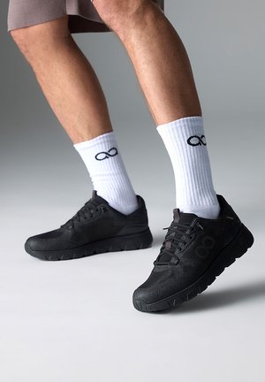 Sorte atletiske sneakers med mesh-overdel, tekstureret overlay og tykke såler. Bruges sammen med hvide ribbede sokker med et sort uendelighedssymbol.