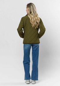 Olivovo zelená prešívaná bunda s hnedým golierom, v kombinácii s svetlomodrými džínsami a bielymi teniskami. Dizajn obsahuje diamantový vzor.