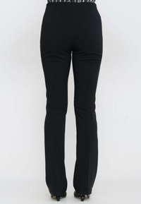 Donna che indossa pantaloni attillati neri e scarpe con tacco nere, in piedi contro uno sfondo bianco semplice, vista da dietro.