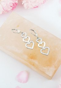 Silverörhängen med tre sammanlänkade hjärtformer, med en glittrande kant av klara stenar. Mot en ljus stenbakgrund.