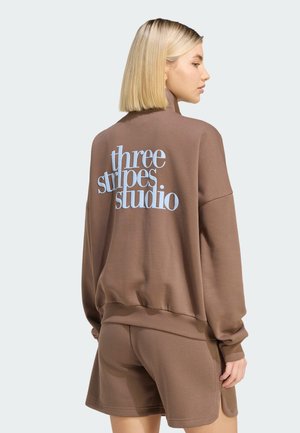Kvinde med blond bob-frisure iført brun sweatshirt og matchende shorts, ryggen vendt mod kameraet, med teksten "three stripes studio" på sweatshirt.