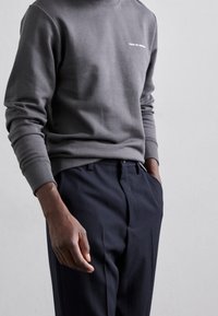 Gray sweatshirt with a crew neck and ribbed cuffs. Features white text on the chest. Paired with navy trousers, showcasing a relaxed fit.