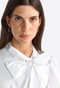 Weiße Bluse mit großem Schleifenkragen, strukturiertem Stoff und langen Ärmeln; das Modell hat lange dunkle Haare und trägt Ohrringe in Form von Ringen.
