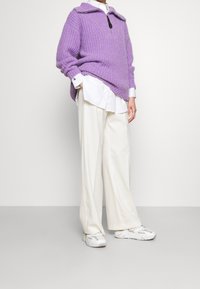 Pull-over tricoté violet avec col à fermeture éclair, porté sur une chemise blanche. Pantalon blanc ample et baskets gris clair complètent la tenue.