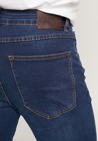 Mörkblå denimjeans med en läderlapp vid midjan. Funktioner inkluderar en bakficka med synlig sömdetalj.