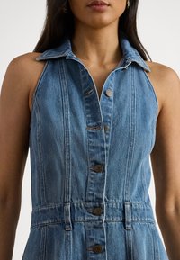 Džinsa kleita ar apkaklīti, bez piedurknēm, gaiši zilā krāsā, ar rāvējslēdzēja aizdari priekšpusē un redzamām šuvju detaļām.