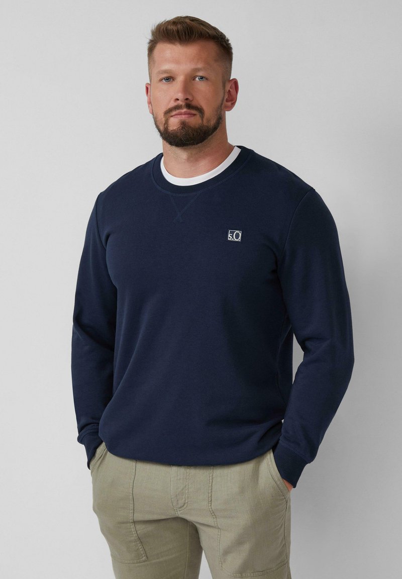 Uomo con barba corta che indossa un felpa blu navy e pantaloni beige, in piedi con le mani in tasca su uno sfondo grigio chiaro.