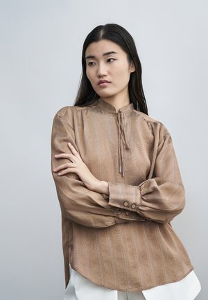 Mlada žena s dugom tamnom kosom nosi svijetlosmeđu prugastu bluzu s kravatom na vratu i bijele hlače, stoji s prekriženim rukama.