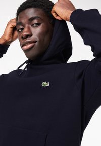 Sweat à capuche bleu marine en tissu doux avec une poche kangourou, arborant un petit logo crocodile Lacoste vert et blanc sur la poitrine.