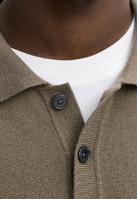Brauner Strickcardigan mit V-Ausschnitt, darunter ein kontrastierendes weißes T-Shirt und zwei schwarze Knöpfe am Kragen. Weicher Stoff, keine Muster.