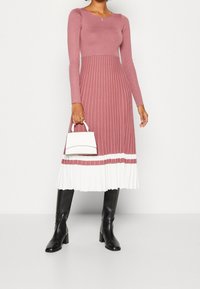 Pitkähihaisen, muotoilultaan istuvan pehmeän vaalenpunaisen mekon ribbitekstuuri, joka siirtyy valkoisiin laskoksiin helman kohdalla. Parina valkoinen muotoiltu käsilaukku ja mustat saappaat.