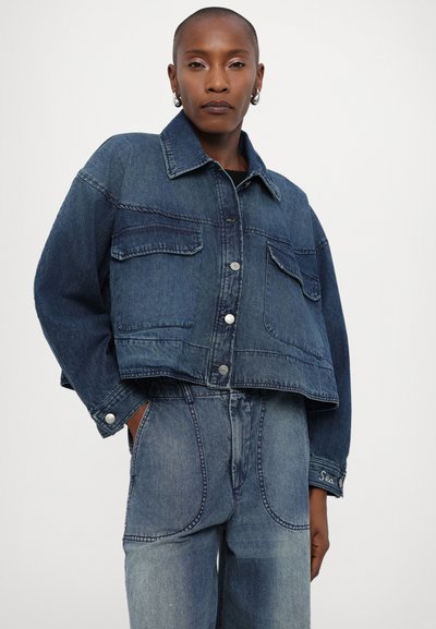 Denim jakna s kročenim dizajnom, temno modre barve, dvema žepoma na prsih, srebrnimi gumbi in širokimi rokavi, v kombinaciji z ustreznimi denim hlačami.