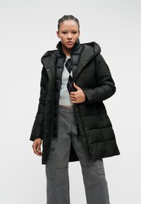 Sort pufferjakke med hætte, med en quiltet design, frontlynlås og trykknapper, båret over en grå argyle sweater og grå bukser.