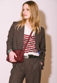 Femme avec de longs cheveux blonds portant un costume marron, un t-shirt graphique rayé, un bracelet turquoise et tenant un sac bandoulière bordeaux.