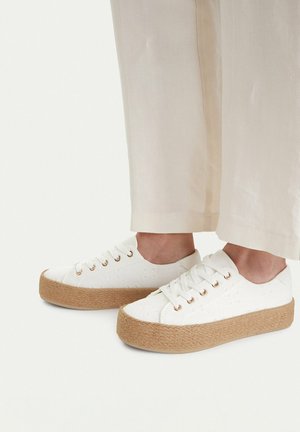 Füße in weißen Canvas-Plattform-Sneakern mit dicken, geflochtenen beigen Sohlen und weißen Schnürsenkeln, kombiniert mit elfenbeinfarbenen Hosen.