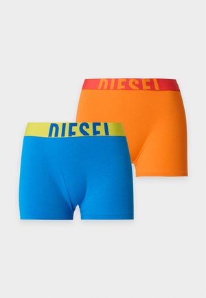 Femme portant un soutien-gorge de sport blanc et un short boxer orange avec des bandes rouges à la taille, tous deux estampillés du mot Diesel, debout pieds nus sur fond blanc.