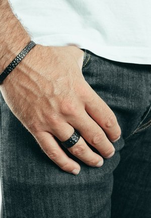 Black textured ring and matching black leather bracelet on a hand, with visible skin texture and fingers against dark jeans.