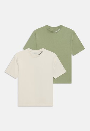 Deux t-shirts à col rond pliés, un beige devant et un vert olive derrière, tous deux avec un petit logo Calvin Klein près de l'encolure.