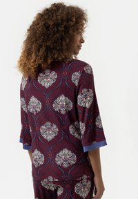 Bordeauxgekleurde top met bloemenpatronen, driekwartmouwen en blauwe manchetten. Zacht materiaal met intricate details en textuur.