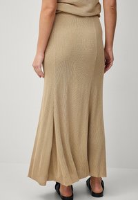 Maxi nederdel i beige ribbet stof med en sideslids for ekstra bevægelse, parret med sorte flade sko, der viser en glat, struktureret overflade.