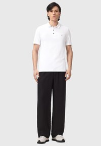 Giovane uomo in piedi, che indossa una polo bianca a maniche corte, pantaloni neri a gamba larga e sneakers bianche, su uno sfondo chiaro e semplice.