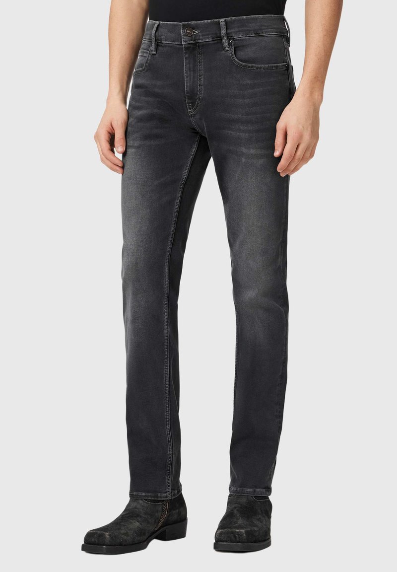 Mies pukeutunut slim fit -mallisiin tummanharmaisiin farkkuihin ja mustiin mokkanahkasaappaisiin, seisoo yksinkertaisen vaaleanharmaan taustan edessä.
