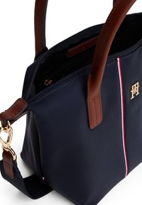 Marineblauwe nylon handtas met bruine leren accenten, voorzien van een ritsluiting, binnenzakken en een gouden logo met witte en rode strepen.