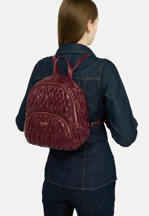 Kvinde i mørk denimjakke og jeans, der bærer en kviltet bordeaux læderrygsæk med guld lynlåse og logo på ryggen.