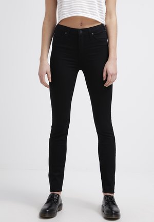 Femme portant un jean noir ajusté, un top blanc rayé court et des chaussures noires à lacets, debout sur un fond clair uni.