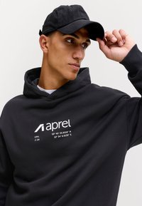 Mladi moški, oblečen v črno kapuco z belim besedilom in koordinatami, prilagaja črno kapo na preprostem svetlem ozadju.