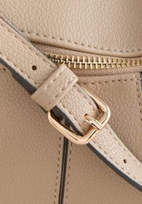 Correa para bolso de cuero beige con una hebilla de metal dorado, presenta una superficie texturizada y costuras visibles cerca de una cremallera.