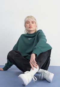 Zelena dukserica s visokim izrezom i Nike logom, crne traperice opuštenog kroja, bijele tenisice, sjedeći položaj na plavoj površini, neutralna pozadina.