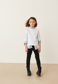 Bambino piccolo con capelli a spalla in piedi, indossa una felpa bianca sopra una camicia blu, pantaloni neri e stivali neri con lacci su uno sfondo semplice.