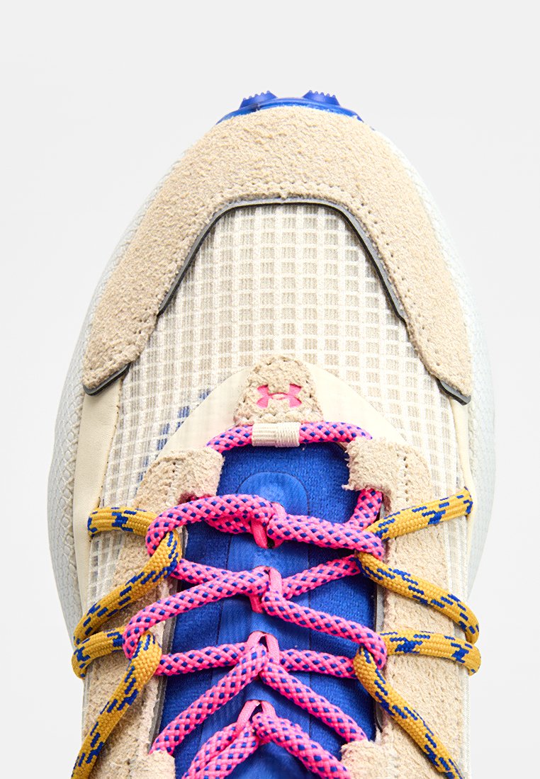 El zapato presenta una parte superior de textil beige con un patrón en cuadrícula, acentos azules y cordones vibrantes en rosa y amarillo, con una puntera de ante texturizada.