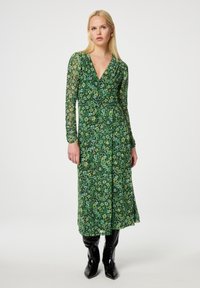 Grünes, blumiges Wickelkleid mit langen, transparenten Ärmeln, V-Ausschnitt, fließendem Rock und mehrlagiger Textur. Tragen mit schwarzen kniehohen Stiefeln.