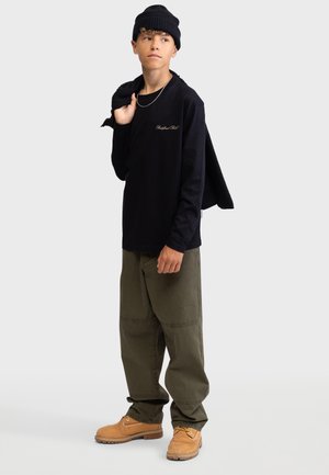 Chemise noire à manches longues avec logo brodé, pantalon baggy vert olive, bottes beige avec lacets, et un bonnet en tricot noir. Pull drapé sur l'épaule.