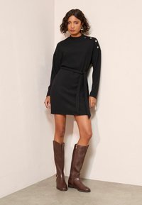 Pletené černé šaty s páskem v pase, žebrovanou texturou a knoflíkovým detailem na rameni. V kombinaci s hnědými koženými botami nad kolena.