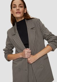 Blazer gris à rayures avec un tissu léger, deux boutons, revers à crans et manches retroussées, porté par-dessus un top noir à côtes.