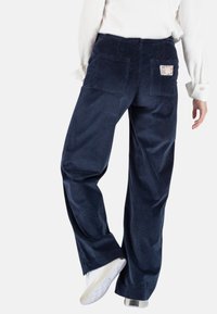Marineblauwe corduroy wijde broek met achterzakken en een gemusteret label. Gladde textuur met verticale ribbels. Gestyled met witte sneakers.