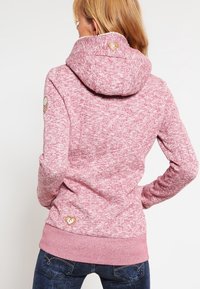 Fleecepaita vaaleanpunaisessa marling-kuviossa ja korkeassa kauluksessa. Takanapaita on varustettu logolla ja hihansuiden peukalonauhoilla.