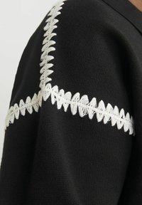 Îmbrăcăminte neagră cu un broderie albă în zig-zag proeminentă pe cusătura umărului, evidențiind textura și contrastul materialelor.
