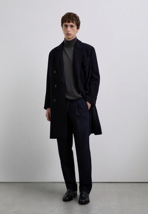 Abrigo azul marino de doble botonadura con grandes botones sobre un suéter gris de cuello alto, combinado con pantalones oscuros de corte entallado y mocasines negros sobre un fondo neutral.