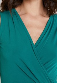 Teal wrap-top i slätt tyg, med en djup V-ringning, samlade detaljer vid axlarna och en mjuk drapering.