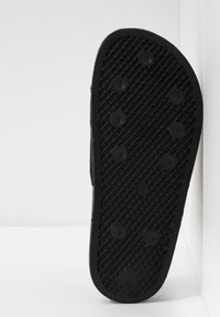 Semelle en caoutchouc noire d'une chaussure avec des motifs circulaires antidérapants et un design en grille texturée, positionnée verticalement sur un fond blanc.