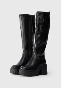 Μαύρες μπότες μέχρι το γόνατο από λεία συνθετική δερματίνη. Παχύ μπλοκ τακούνι, στρογγυλή μύτη, ολοκληρωμένες με πλάγιους φερμουάρ και λεπτομέρειες ραψίματος.