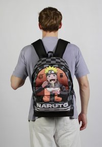 Rugzak met een dambordpatroon en een kleurrijk animekarakter, voornamelijk in oranje en zwart, die een kom noedels vasthoudt.