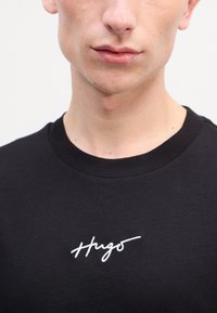 Czarna bawełniana koszulka z okrągłym dekoltem, z białym haftowanym logo "Hugo" na przodzie. Gładka tekstura, dopasowany krój.