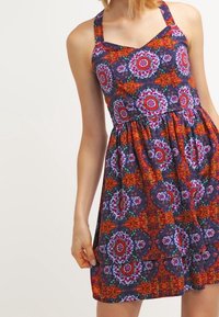 Robe imprimée florale avec un corsage ajusté, des bretelles larges et une jupe évasée. Les couleurs comprennent des motifs bleu marine, rouge, orange et vert.